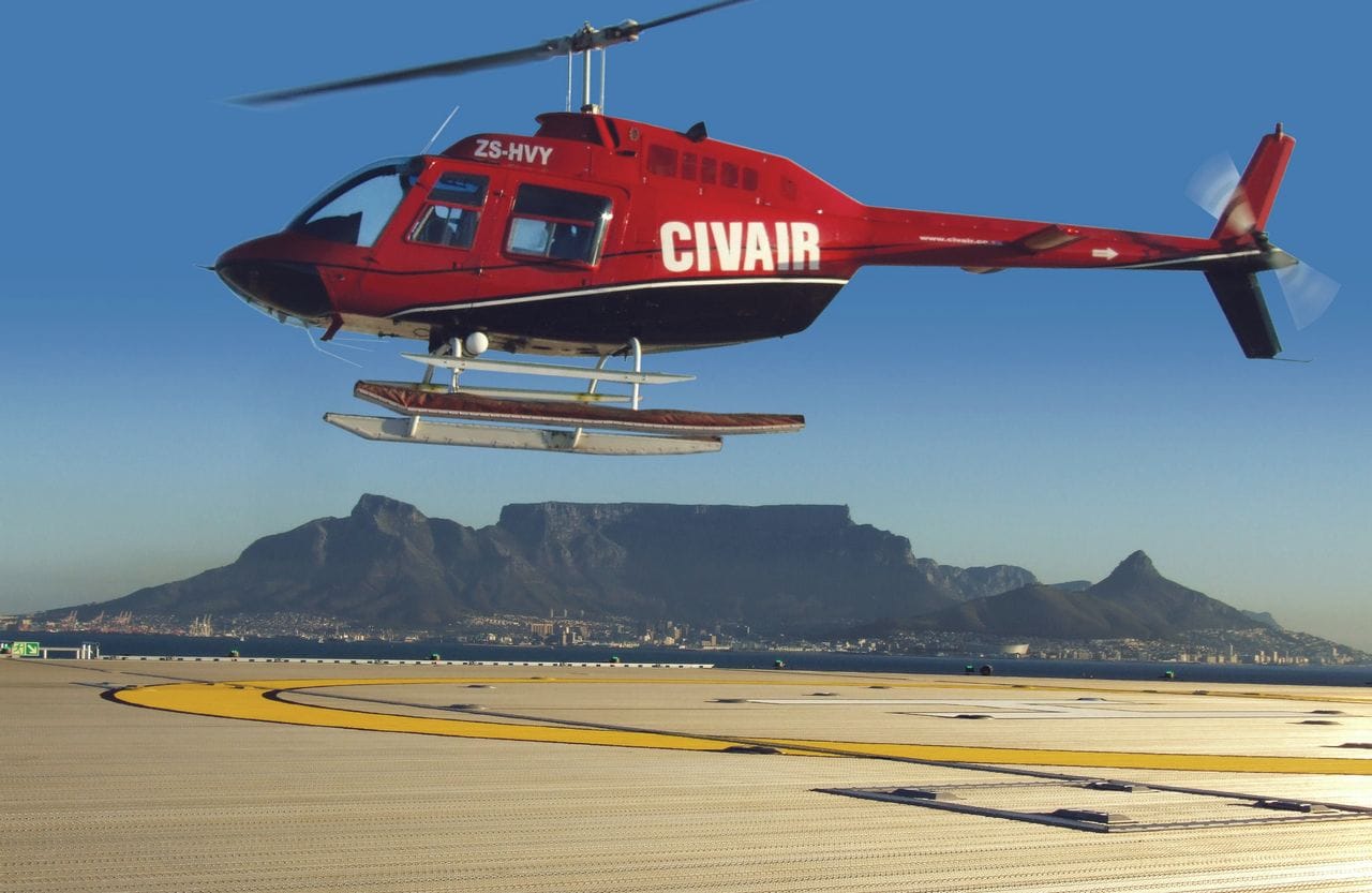CIVAIR’s red Bell 206 JetRanger ZS-HVY lifting off, Table Mountain across the bay behind it